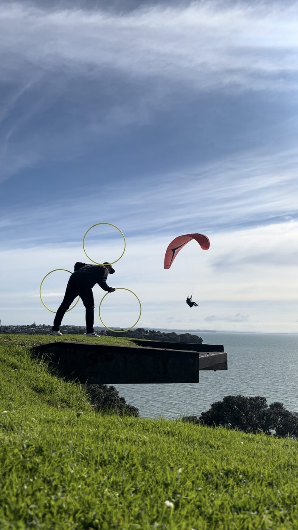 The Catching Machine balancing three hoops on the Auckland coastline as a paraglider flies past